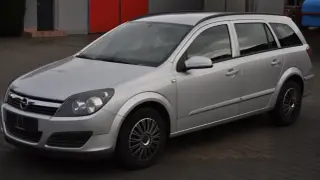 Srebrny Opel Astra H 1.6 benzyna, kombi, zaparkowany na kostce brukowej przed budynkiem z czerwonymi drzwiami.