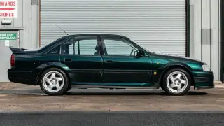 Zielony Opel Omega Lotus z charakterystycznym spojlerem i felgami stoi przed budynkiem z metalową bramą.