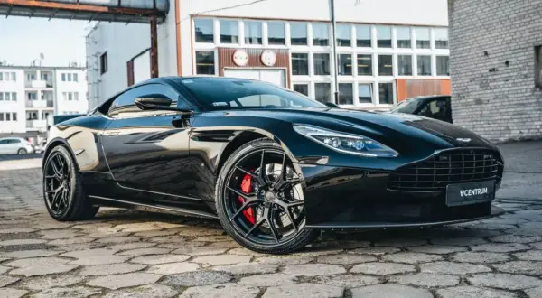 Błyszczący czarny Aston Martin DB11 z czerwonymi zaciskami hamulcowymi na stylowych felgach, zaparkowany na brukowanej ulicy.