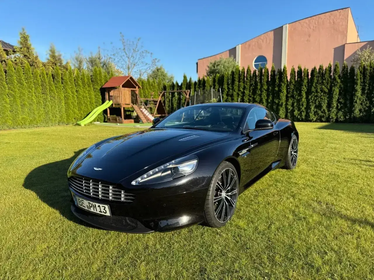 Czarny Aston Martin DBS lśni na trawniku przed domem. W tle plac zabaw i wysokie tuje.