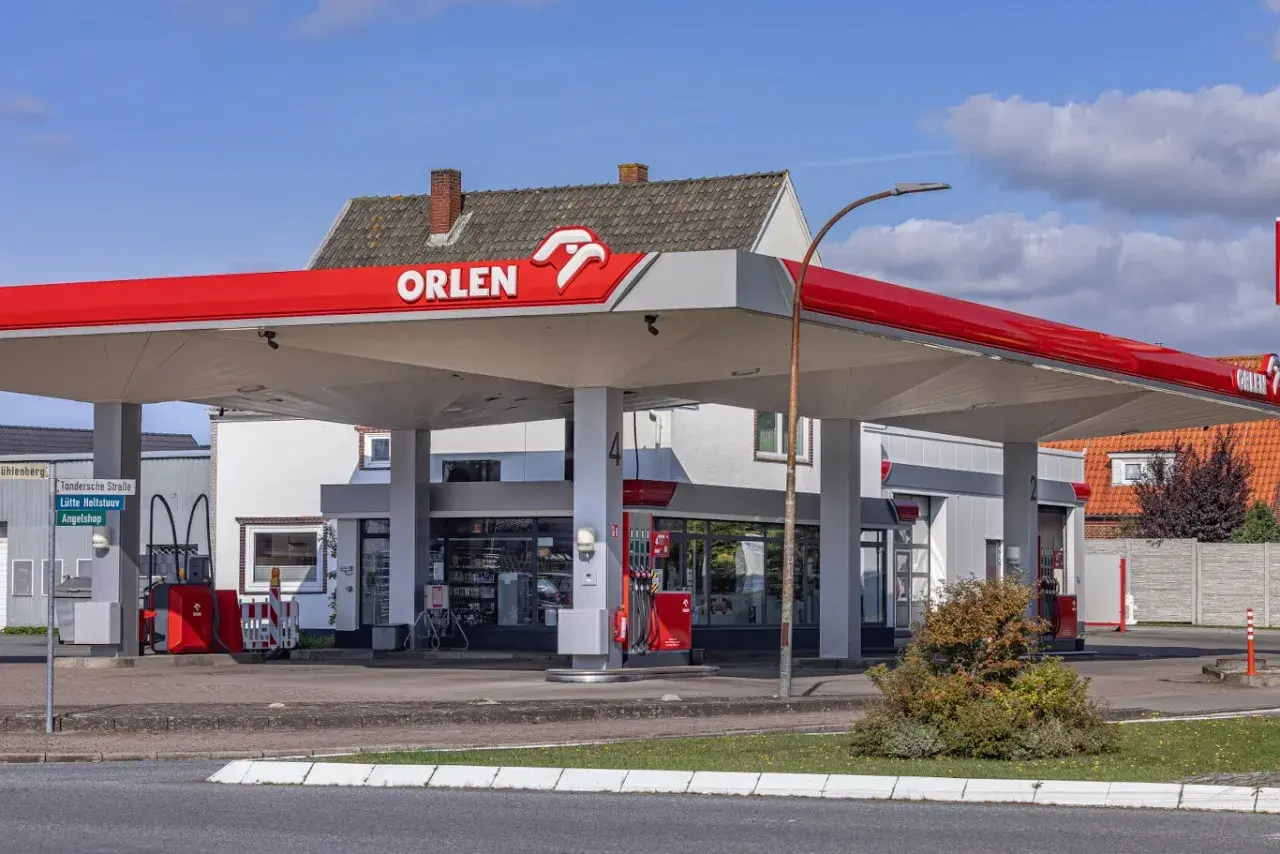 Stacja paliw Orlen z charakterystycznym czerwonym zadaszeniem. W tle budynek z oknami i sklepem.