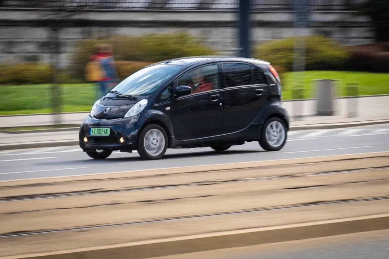Czarny Peugeot iOn jedzie ulicą, w tle rozmyta postać przechodnia.