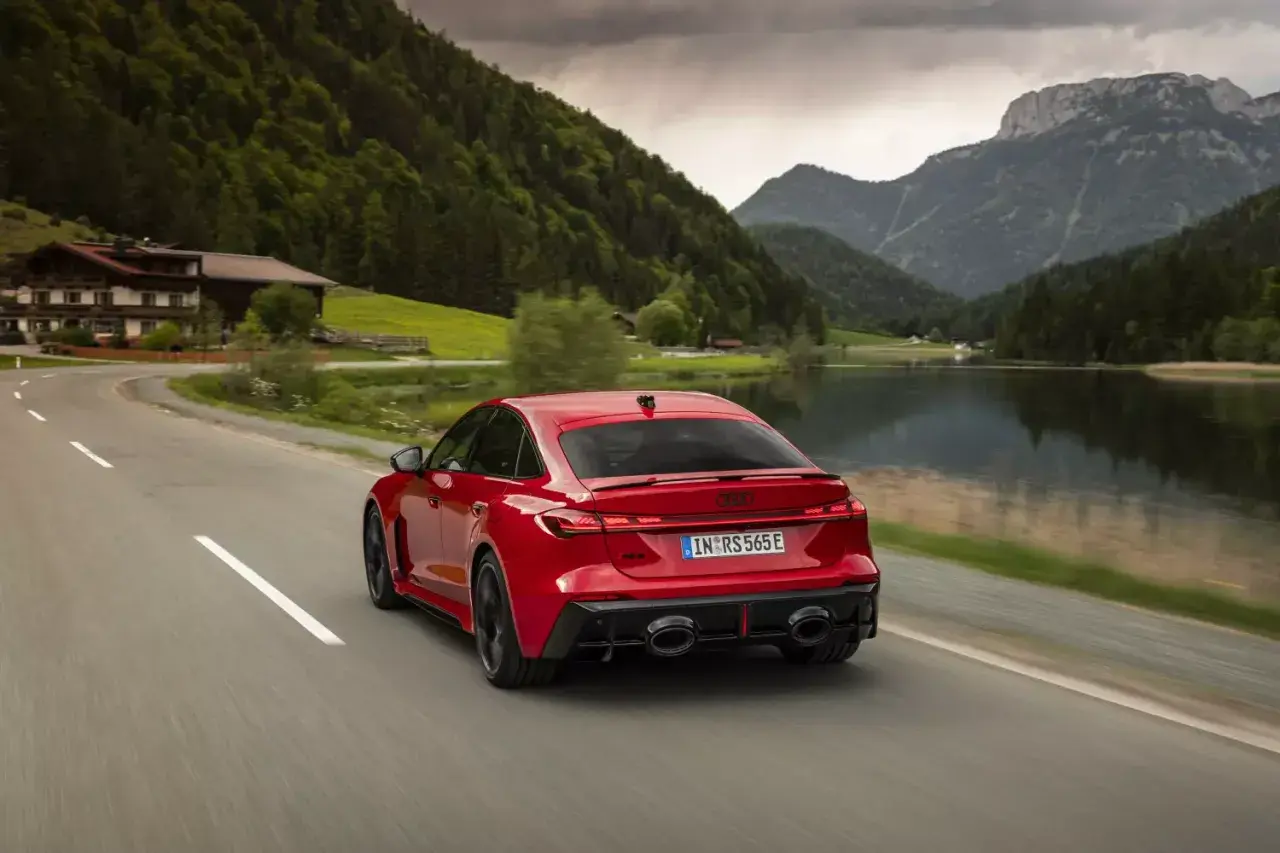 Czerwone Audi RS5 2026 pędzi malowniczą drogą nad jeziorem, otoczone zielonymi wzgórzami i górami.
