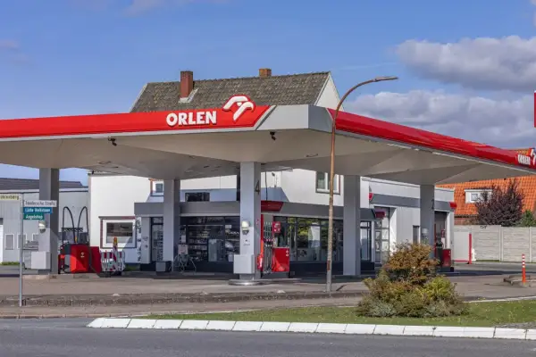 Stacja paliw Orlen z charakterystycznym czerwonym zadaszeniem. W tle budynek z oknami i sklepem.